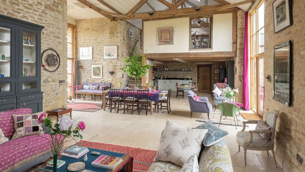 A spacious living area with a high ceiling, exposed wooden beams, a dining area, and a kitchen visible in the background. - Spacious and beautifully converted 18th century stone barn.  Pet-friendly! (Lullington)