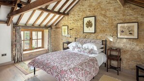 A bedroom with a stone wall, wooden beams, a bed with floral bedding, a nightstand with a lamp, and a window with curtains. - Spacious and beautifully converted 18th century stone barn.  Pet-friendly! (Lullington)