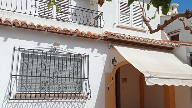 Un patio con una mesa y sillas, un edificio blanco con techo de tejas y un árbol de hojas verdes.