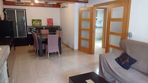 Una sala de estar con sofá, una mesa de centro, una zona de comedor con mesa y sillas, y una cocina visible a través de una puerta abierta.