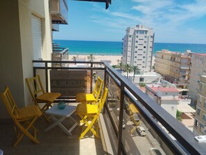 Varanda com cadeiras amarelas e uma mesa, com vista para uma praia e um prédio alto.