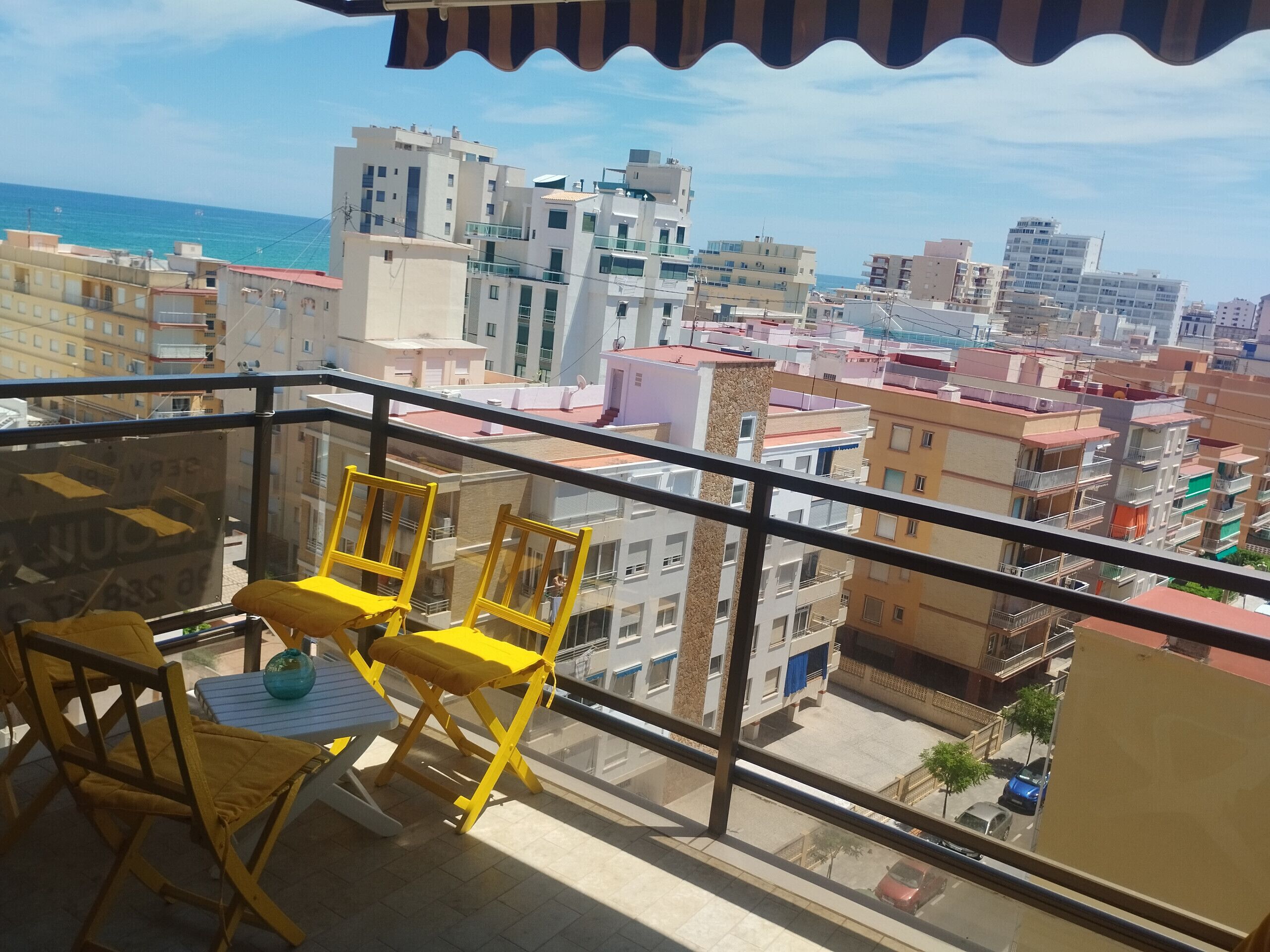 A balcony with yellow chairs and a table, overlooking a cityscape with various buildings and a clear sky.