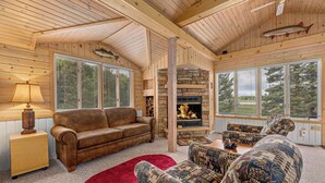 Interior acogedor de una cabaña con chimenea de piedra, sofá de cuero, sillones estampados y ventanas amplias con vistas al exterior.