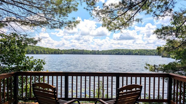 Una terraza de madera con dos mecedoras y una pequeña mesa, con vista a un lago y una orilla boscosa.