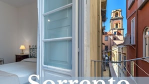 Un dormitorio con cama, mesita de noche y ventana. La habitación tiene vista al centro de la ciudad, con edificios y una torre del reloj.