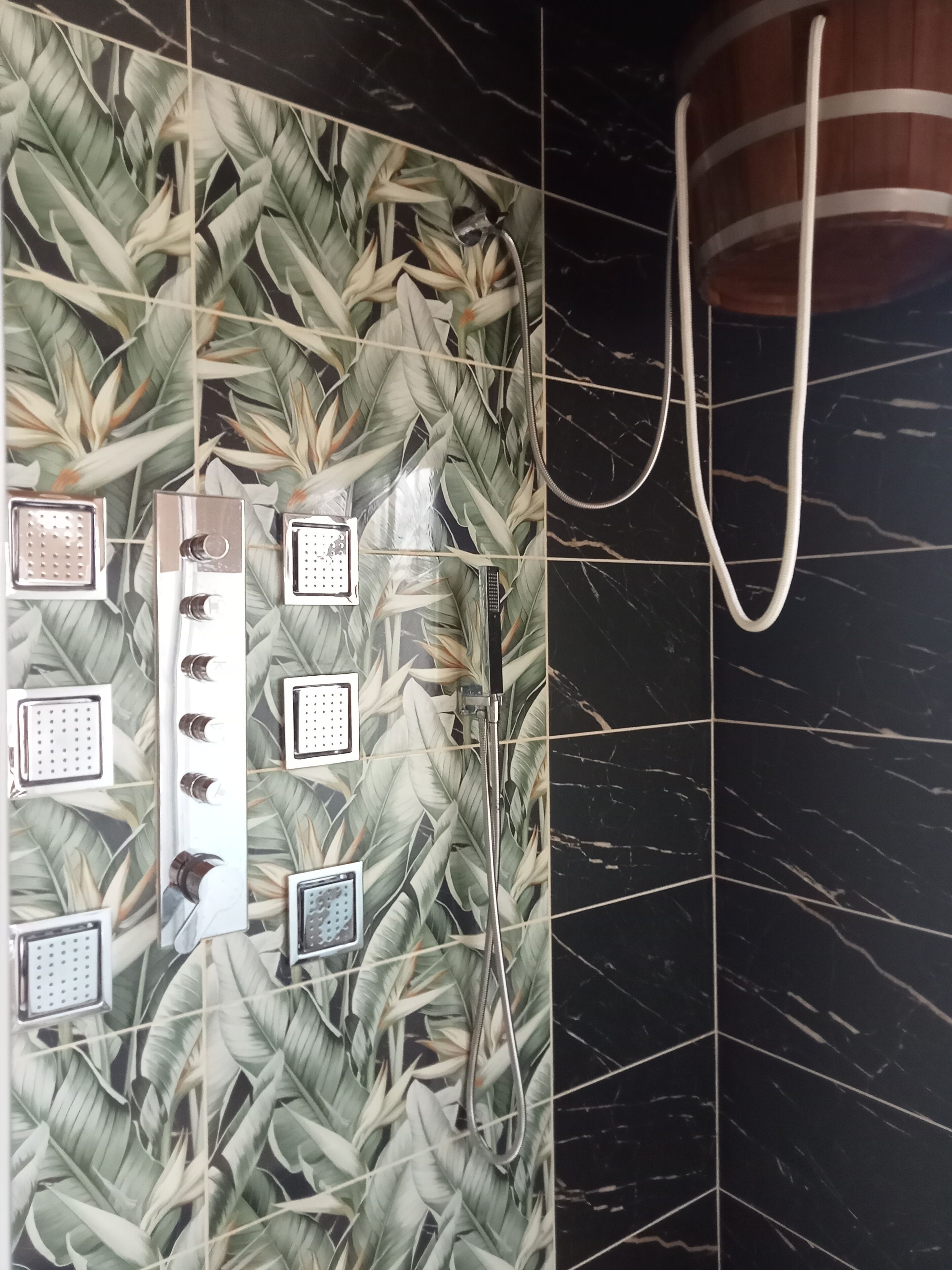 Une salle de bain avec un espace douche agrémenté d’un motif de feuilles tropicales sur le mur, d’un pommeau de douche à main et d’un flexible de douche.
