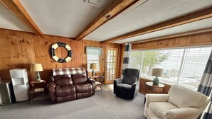 Ein gemütliches Wohnzimmer mit einer braunen Ledersofa, einem schwarzen Sessel und einem weißen Sessel. Auf Beistelltischen stehen zwei Lampen, an einer Holzwand hängt eine runde Dekoration und es gibt ein Fenster mit Jalousien.
