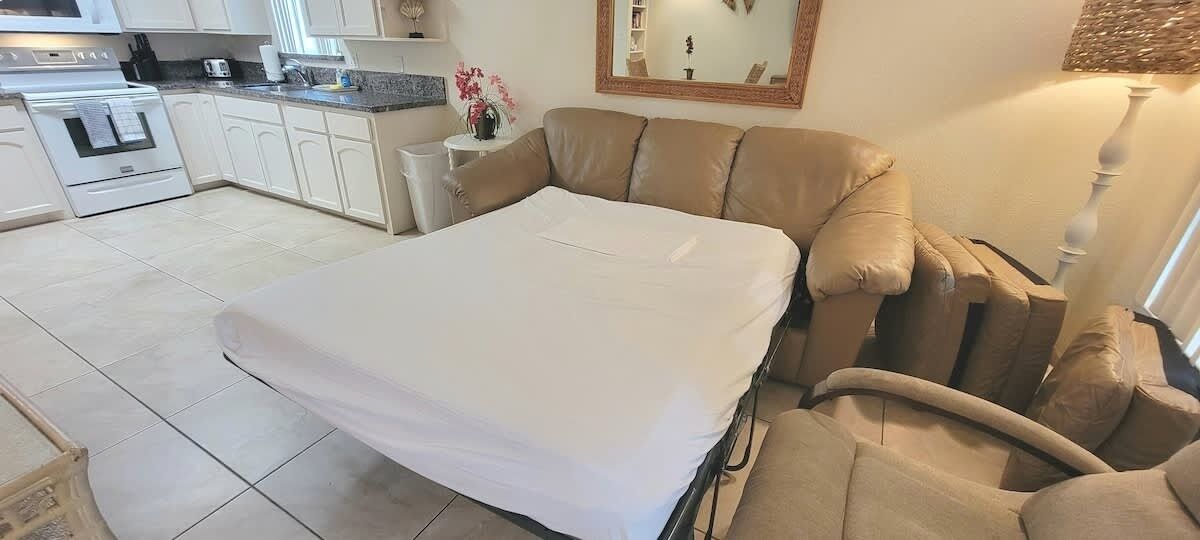 A bedroom with a bed, a sofa, a small table, and a kitchen in the background.