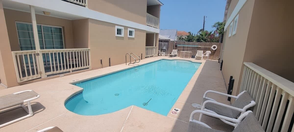 A rectangular swimming pool with a white deck, surrounded by lounge chairs and a small table.