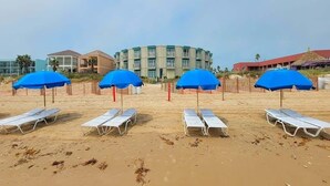 A beach with white lounge chairs and blue umbrellas, a circular building in the background, and a fence separating the beach from the buildings. - Campeche Retreat #12 2 Bedroom Condo (South Padre Island)