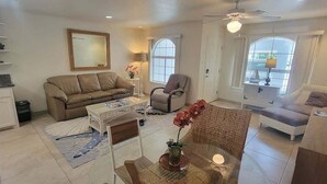 A spacious living room with a sofa, armchair, and coffee table. There is a kitchen area with cabinets and a countertop. The room has large windows and a ceiling fan. - Campeche Retreat #12 2 Bedroom Condo (South Padre Island)