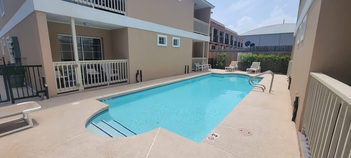 A rectangular swimming pool with a white deck, surrounded by lounge chairs and a building with balconies.