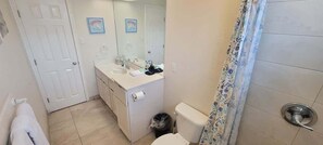 Et badeværelse med toilet, vask og spejl. Der er en dør til venstre og en badeforhæng til højre.