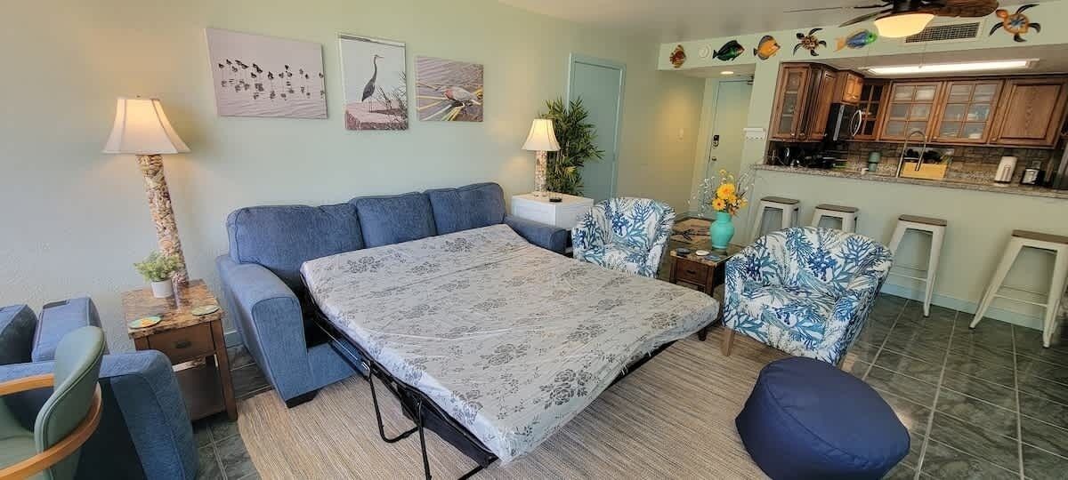 Een woonkamer met een bank, salontafel, fauteuil en een bijzettafeltje met een lamp. Er is een keukenhoek met kasten en een aanrecht.