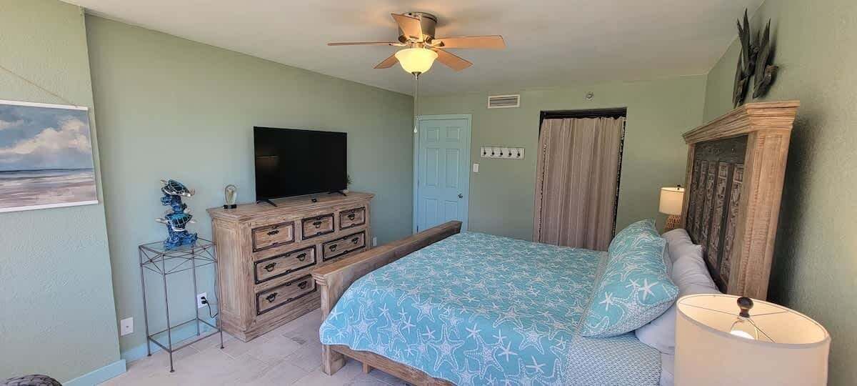 A bedroom with a bed, dresser, television, ceiling fan, and a door.