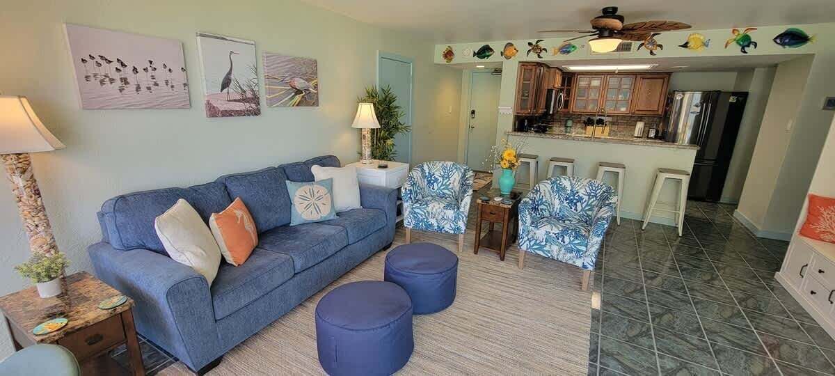 A living room with a sofa, ottomans, and a coffee table. There is a kitchen area with bar stools and a refrigerator.