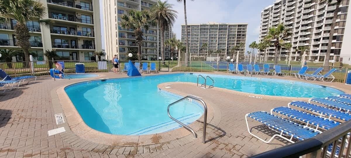 A swimming pool with blue chairs and a lifeguard stand, surrounded by palm trees and high-rise buildings.
