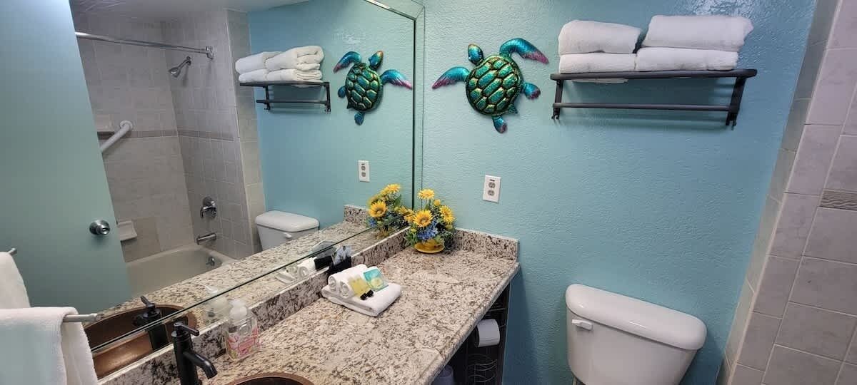 A bathroom with a shower, toilet, and sink. There are towels on a shelf and a mirror with a decorative turtle.