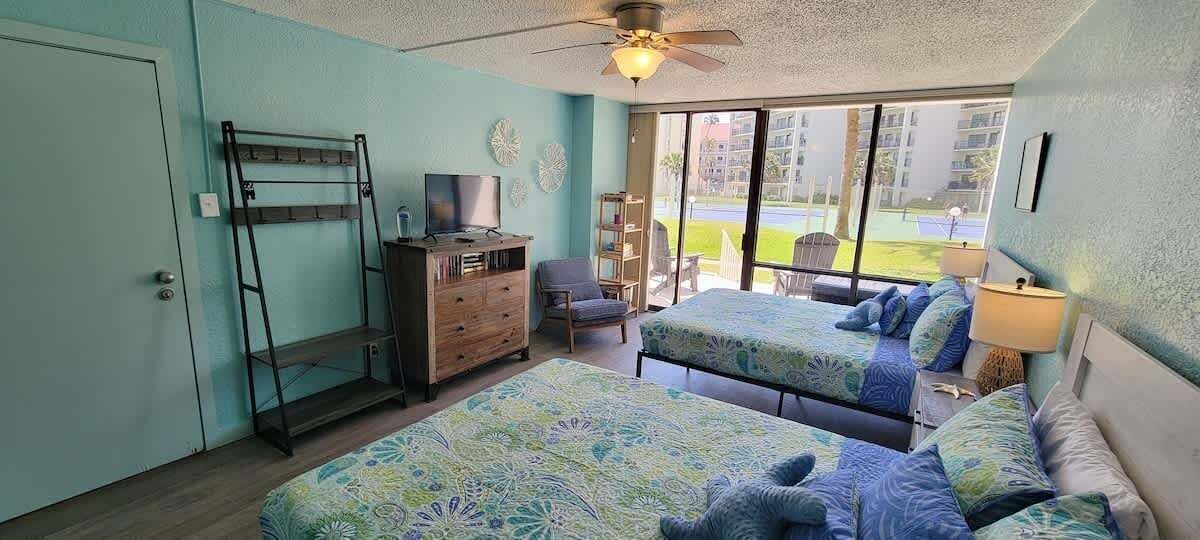 A room with two beds, a TV, a desk, and a chair. There is a balcony with a view of the outside.
