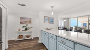 A modern kitchen with light blue cabinets, a white countertop, and a wooden floor. There is a window with a beach view, a hanging light fixture, and a small shelf with baskets. - 🌟Starboard Village 121: Beachfront Corner Unit w/ 2 Balconies🌟 (Pensacola Beach)