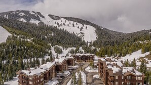 Un complejo turístico en la montaña cubierto de nieve, con varias construcciones tipo chalé, una pista de esquí y un bosque denso.