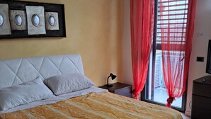 A bedroom with a large bed, a headboard, a bedside table, a lamp, and a window with red curtains. - Home Ross. (Fiorenzuola d'Arda)