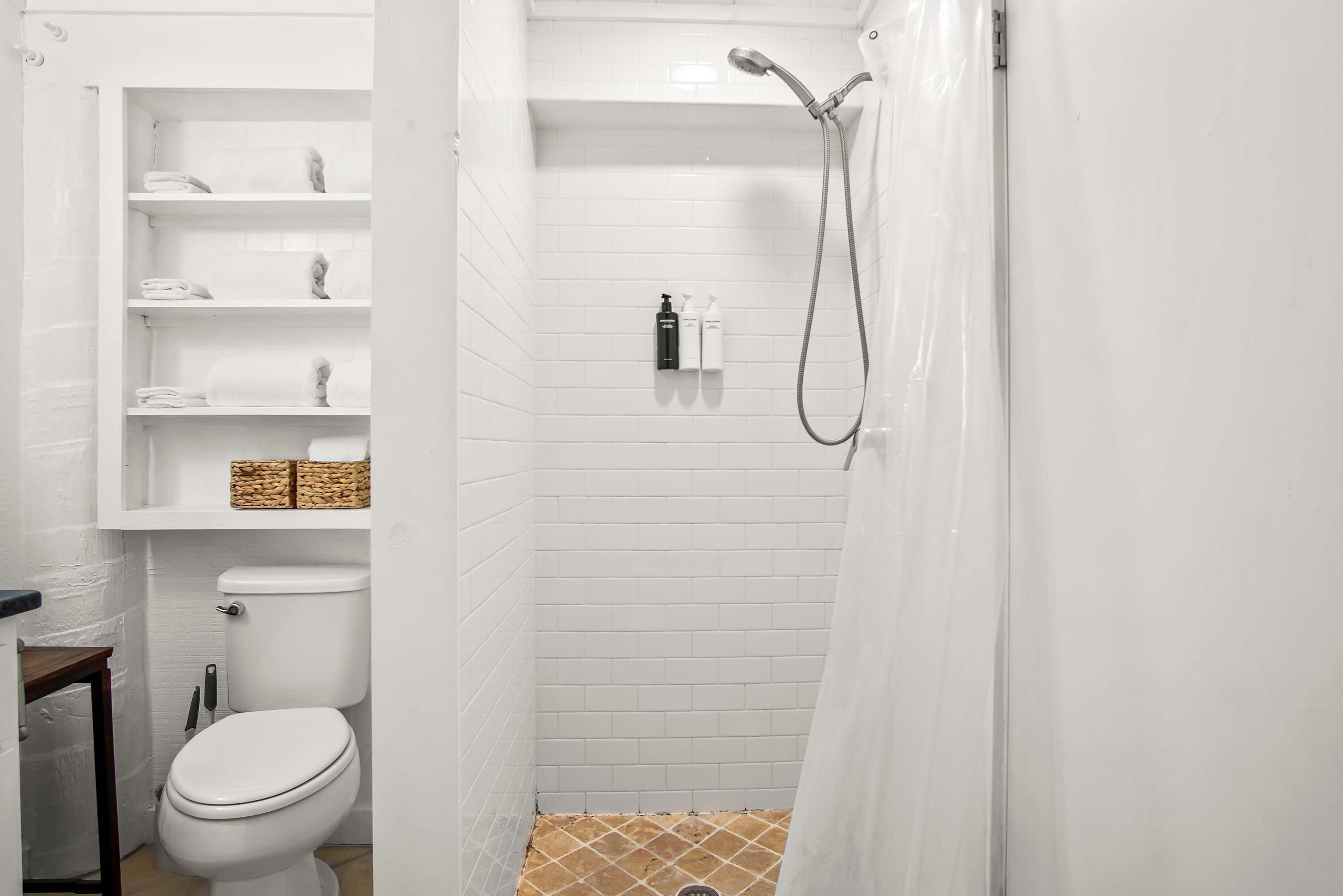 A small, white bathroom with a toilet, shelves with towels, a shower, and a toilet brush.