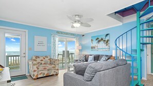 A living room with a grey sofa, a floral armchair, and a turquoise spiral staircase. - Shady Shack West a 3 BR Oceanfront Duplex with Unobstructed Ocean Views (Emerald Isle)