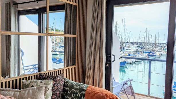Une pièce confortable donnant sur un port de plaisance, avec un canapé agrémenté de coussins, un luminaire en fibres tressées et une grande fenêtre.