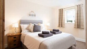 A neatly made bed with pillows and a blanket, a bedside table with a lamp, and a window with curtains. - The Mill set in stunning rural Northumberland (Netherton)