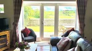 A living room with a sofa, armchair, and a TV on a wooden cabinet. There is a sliding glass door leading to an outdoor area with a table and chairs. - The Mill set in stunning rural Northumberland (Netherton)