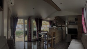 A spacious living area with a sofa, a dining table with chairs, a kitchen area with a TV, and a sliding glass door leading to an outdoor patio.