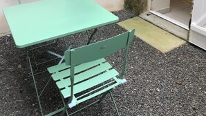 A small outdoor patio with a green metal table and matching chairs, a window, and a stone wall.