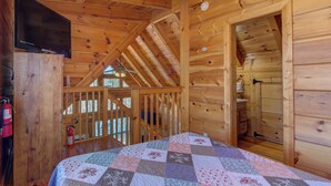 A wooden cabin interior with a bed, a mounted TV, a fire extinguisher, and a staircase leading to an upper level.