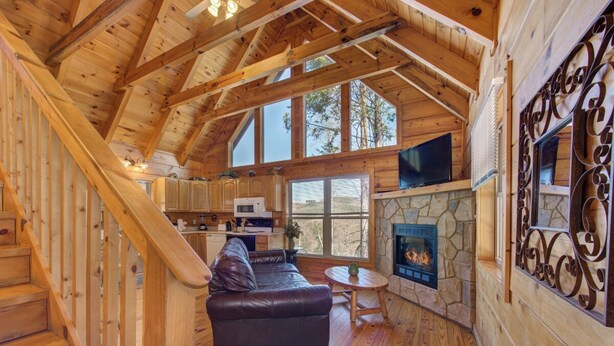 A cozy cabin interior with a stone fireplace, wooden floors, a leather sofa, and a kitchen area.