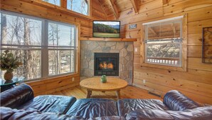 A cozy cabin interior with a stone fireplace, leather seating, and large windows offering views of the surrounding forest.