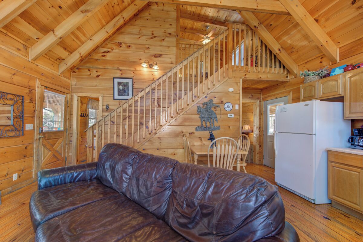 A wooden cabin interior with a leather couch, a staircase, a dining area, and a kitchen.