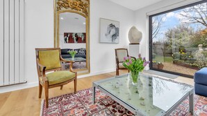 A modern living room with a glass coffee table, two wooden chairs, a patterned rug, a large mirror, and a view of a garden through a large window.