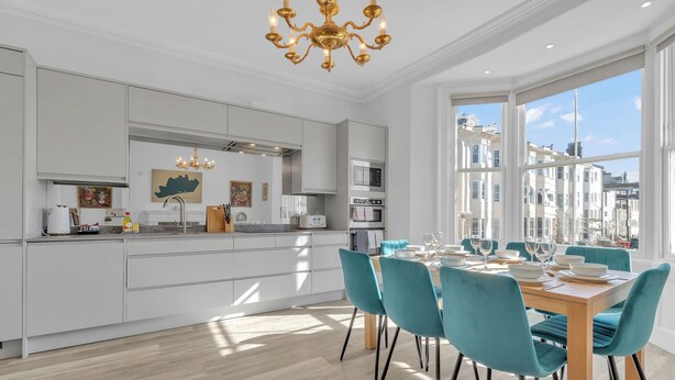 Une cuisine moderne avec des meubles blancs, un coin repas doté de chaises turquoise et une grande fenêtre offrant une vue sur la ville.