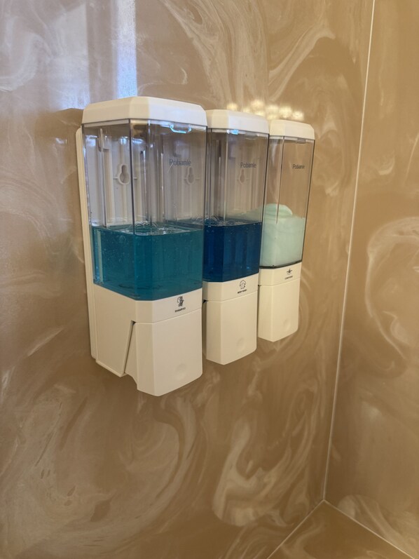 Trois distributeurs contenant des liquides de couleurs différentes, probablement destinés à des produits d’hygiène personnelle, sont fixés à un mur dont la texture ressemble à du marbre.