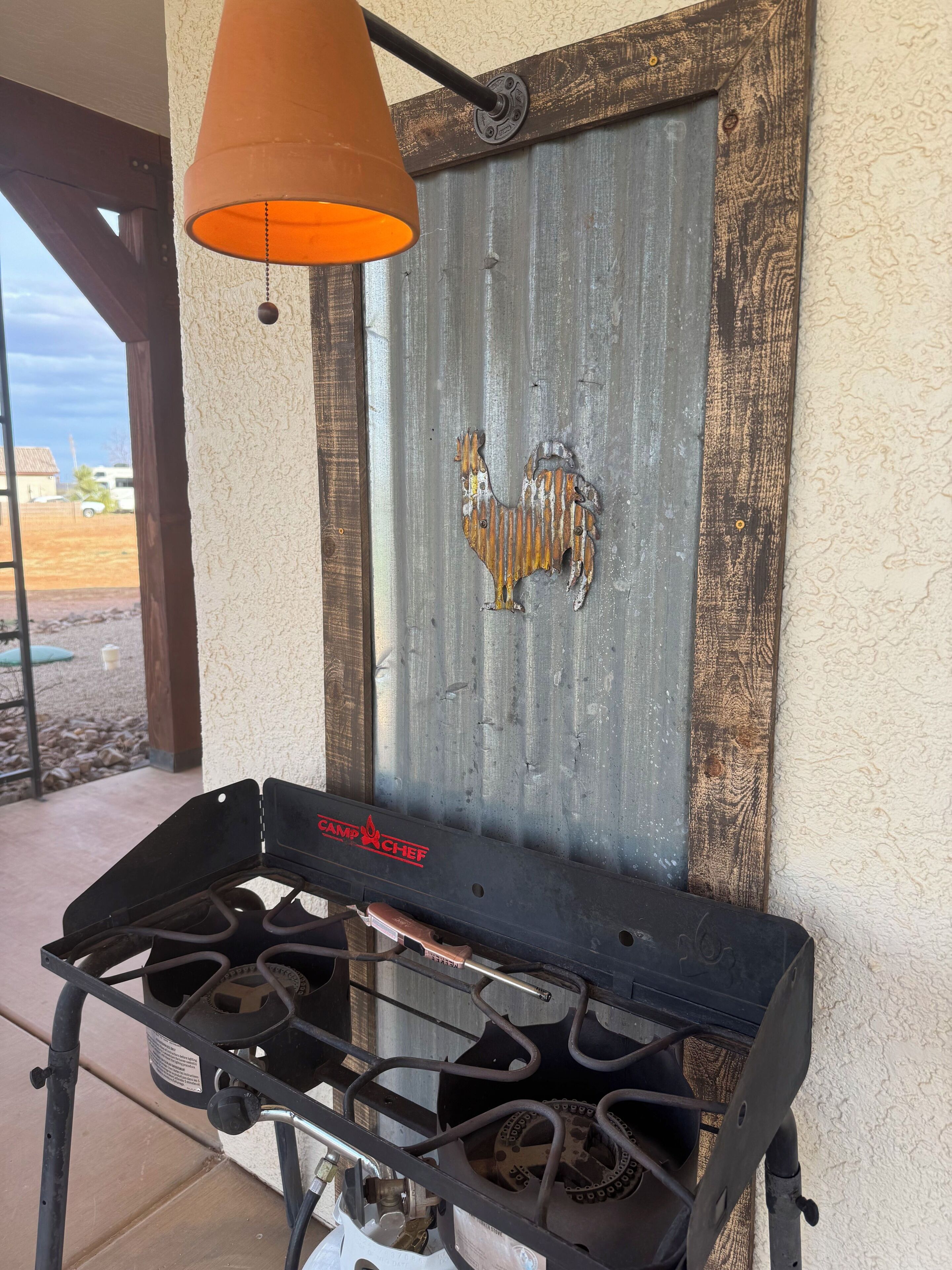 Een draagbaar gasfornuis met een metalen haan decoratie aan een rustieke muur, een hangende oranje lamp en uitzicht op een woongebied.