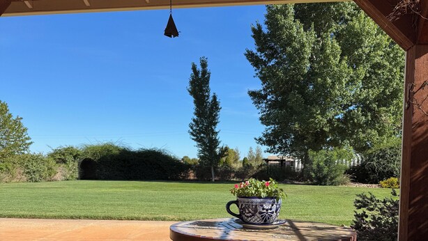 Un patio al aire libre con una mesa redonda y bancos, una lámpara colgante y vistas a un jardín con césped y árboles.