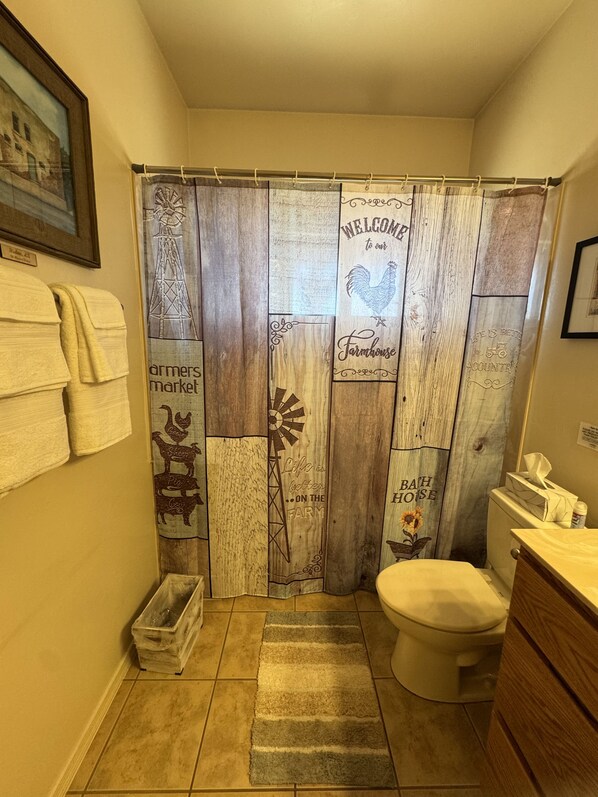 Une salle de bain avec un rideau de douche rustique orné de motifs et d’inscriptions sur le thème de la ferme, des toilettes, une petite poubelle et un porte-serviettes avec des serviettes.