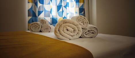 Una cama bien hecha con toallas enrolladas y una cortina estampada al fondo.