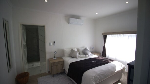 A modern bedroom with a large bed, bedside tables, a nightstand, a wall-mounted air conditioner, and a window with curtains. - Little Bush Guesthouse (Omaruru)