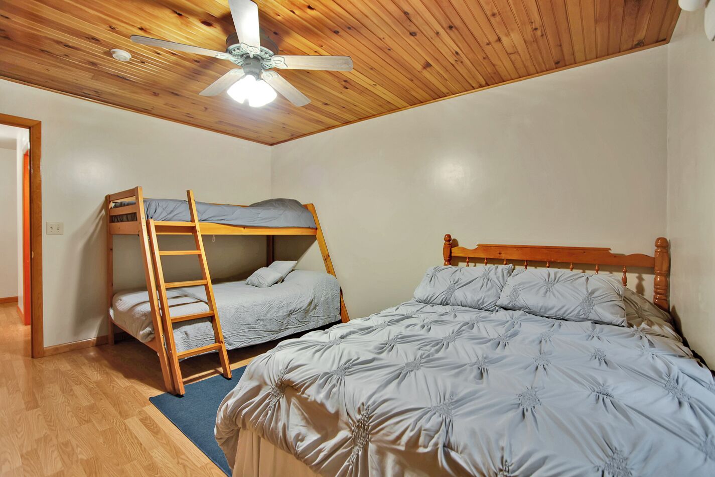 A room with a bunk bed and a single bed, wooden ceiling, and a ceiling fan.