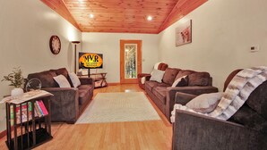 A cozy living room with two brown sofas, a wooden ceiling, a clock, a TV displaying 'MVR Vacation Rentals', and a door leading outside. - Wellsboro Woods Retreat - Near 2 State Parks (Middlebury Township)