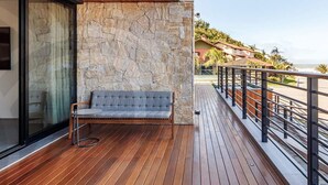 Eine Holzterrasse mit einer Steinmauer, einer grau gepolsterten Bank und Blick auf das Meer und Gebäude im Hintergrund.