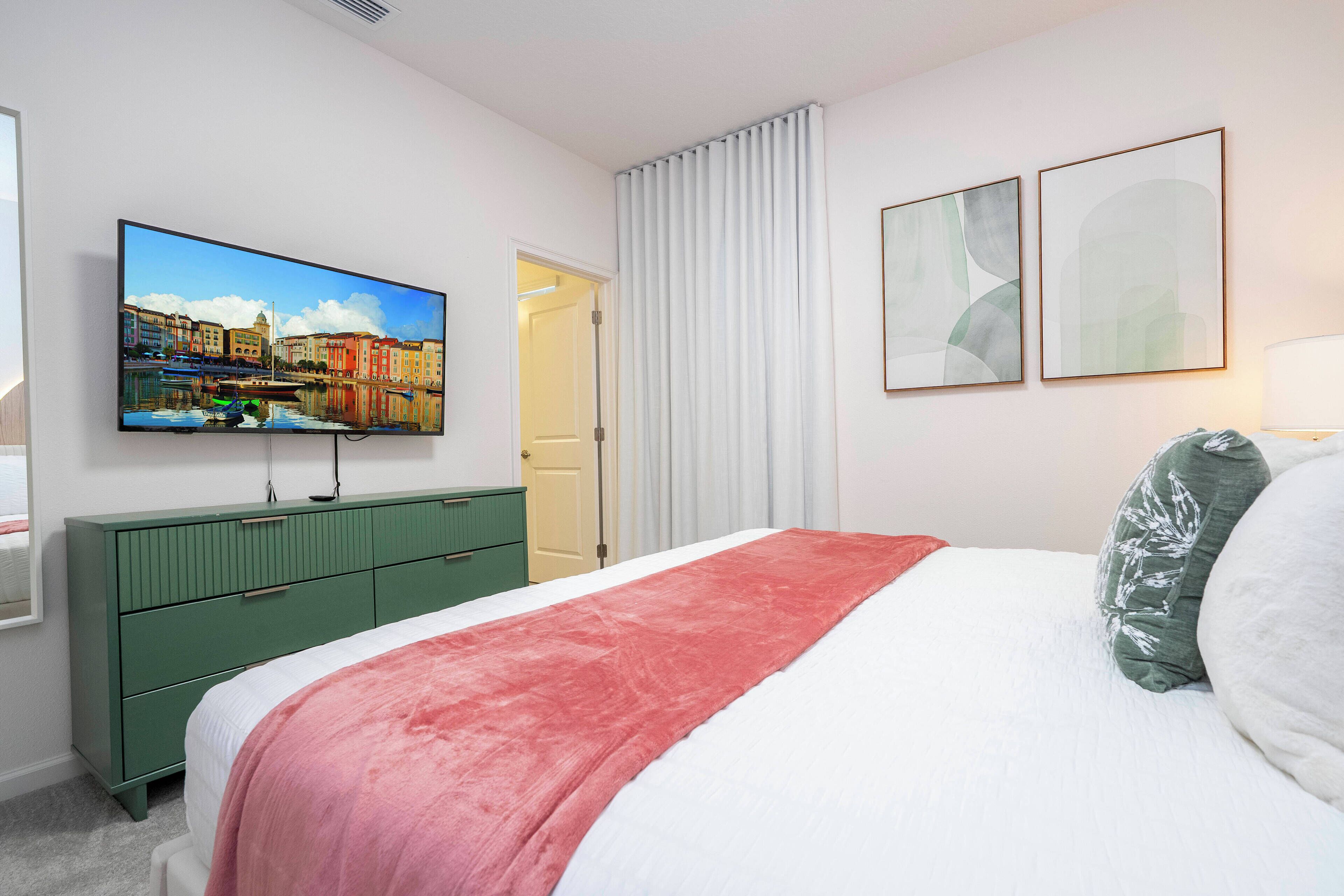 A hotel room with a bed, a green dresser, a television displaying a cityscape, and two abstract paintings on the wall.
