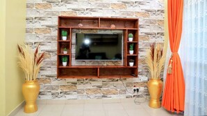 A living room with a stone-patterned wall, a mounted TV, and two golden vases with plants. - Blodriff Apartments (Mombasa)